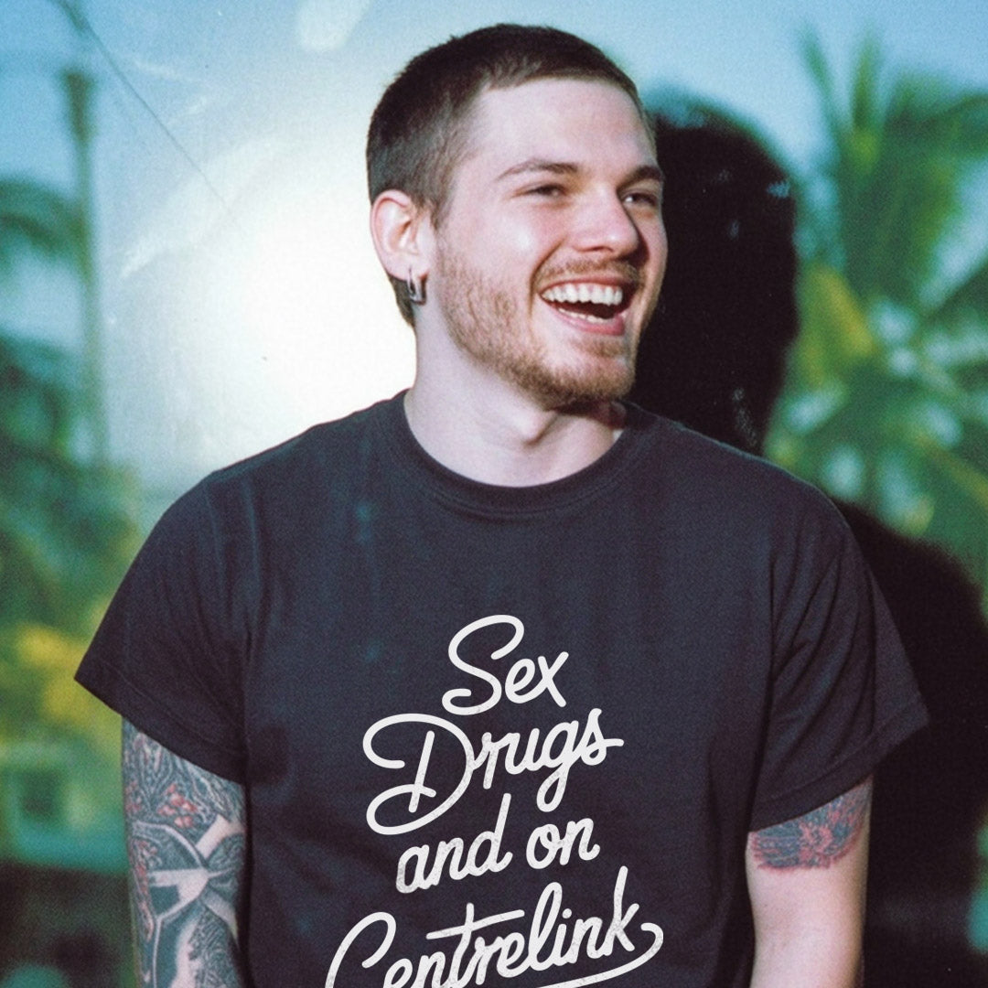 Man wearing a black t-shirt with 'Sex Drugs and on Centrelink' text, standing outdoors with greenery in the background.